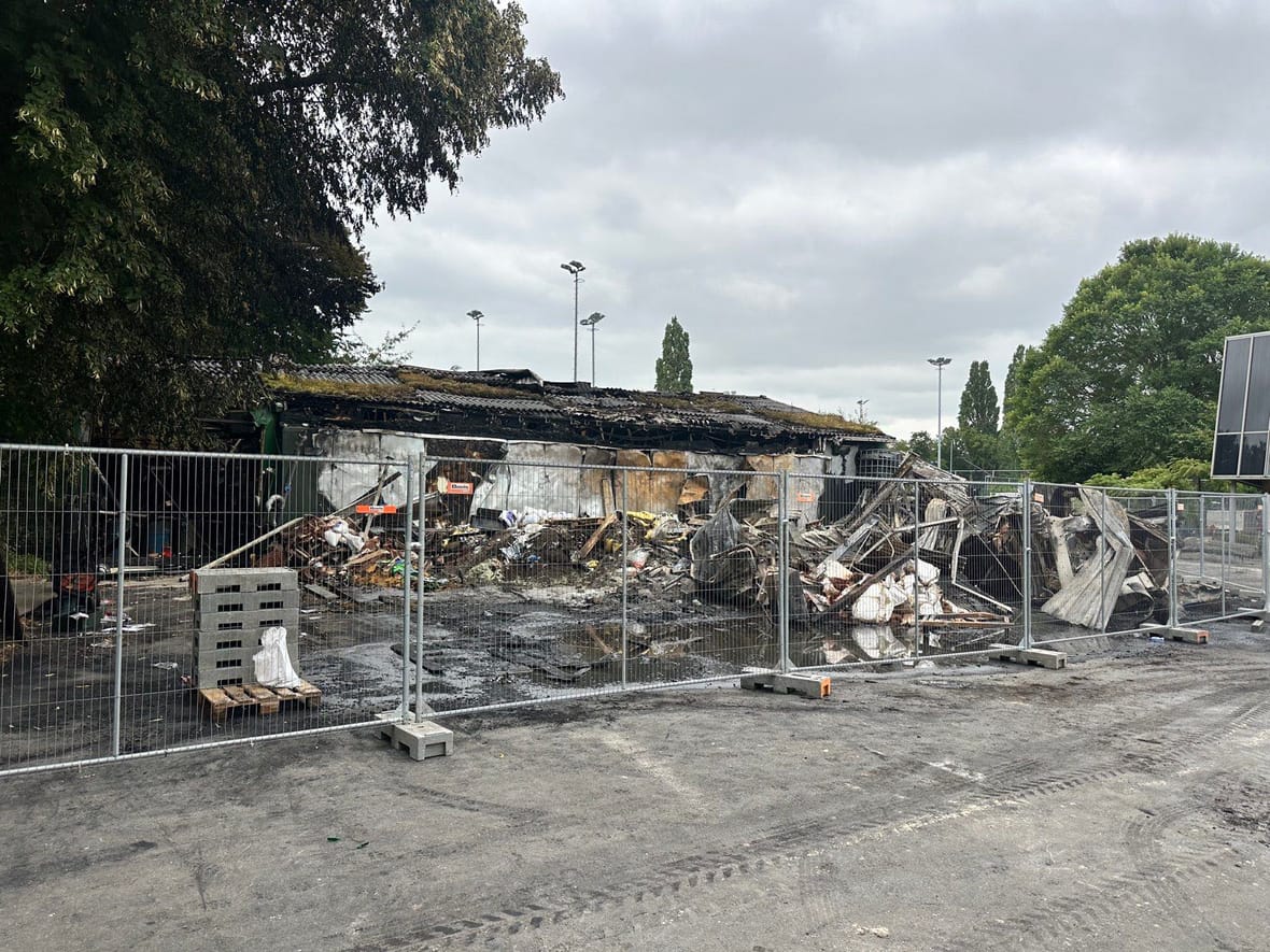 De uitgebrande loods op Sportpark Middenmeer achter bouwhekken, met verwoest materieel en golfplaten op de voorgrond