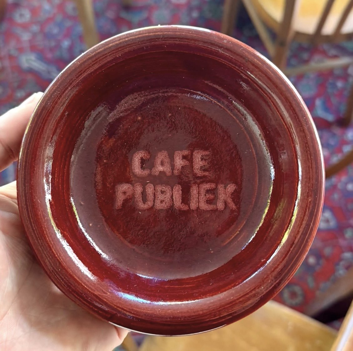 Een dieprode keramische kom met ‘Café Publiek’ in de klei gestempeld, vastgehouden boven een Perzisch tapijt