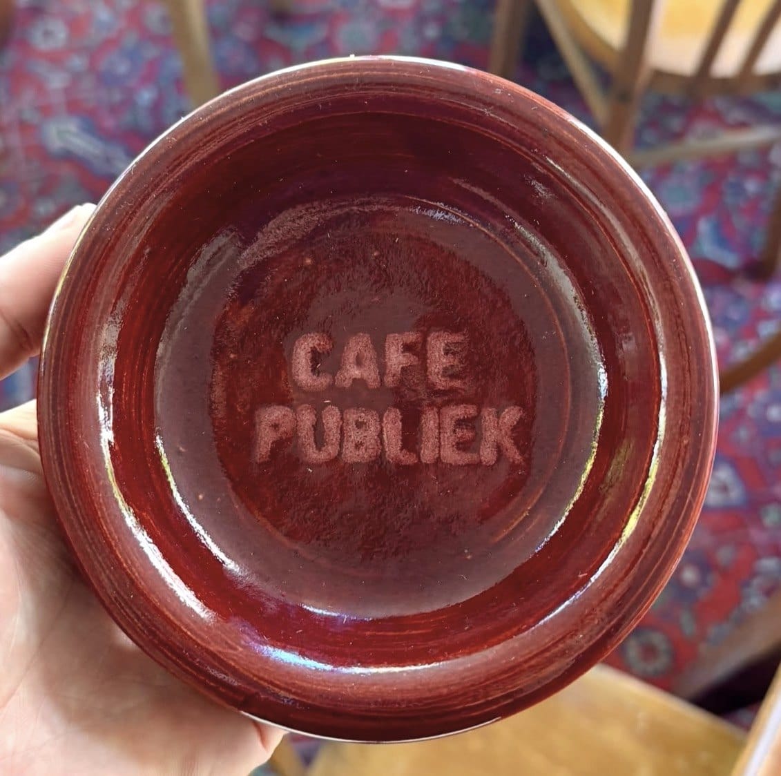 Een dieprode keramische kom met ‘Café Publiek’ in de klei gestempeld, vastgehouden boven een Perzisch tapijt
