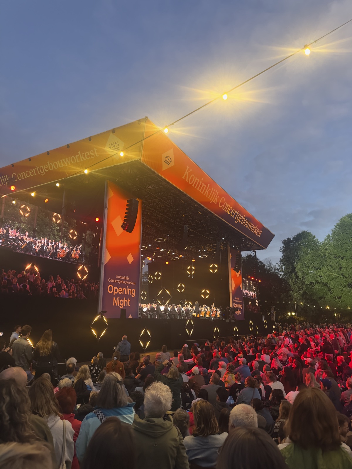 Bezoekers op het gras van het Oosterpark bij schemering, met het verlichte podium op de achtergrond