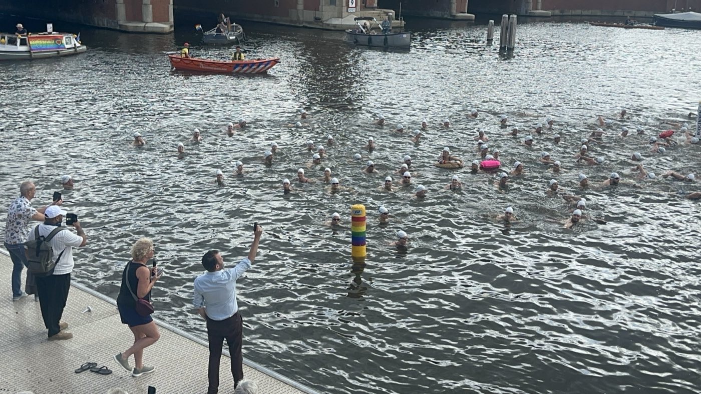 Zwemmers tijdens LoveSwim in de Amstel, met een regenboogboei op de voorgrond en toeschouwers op de kade