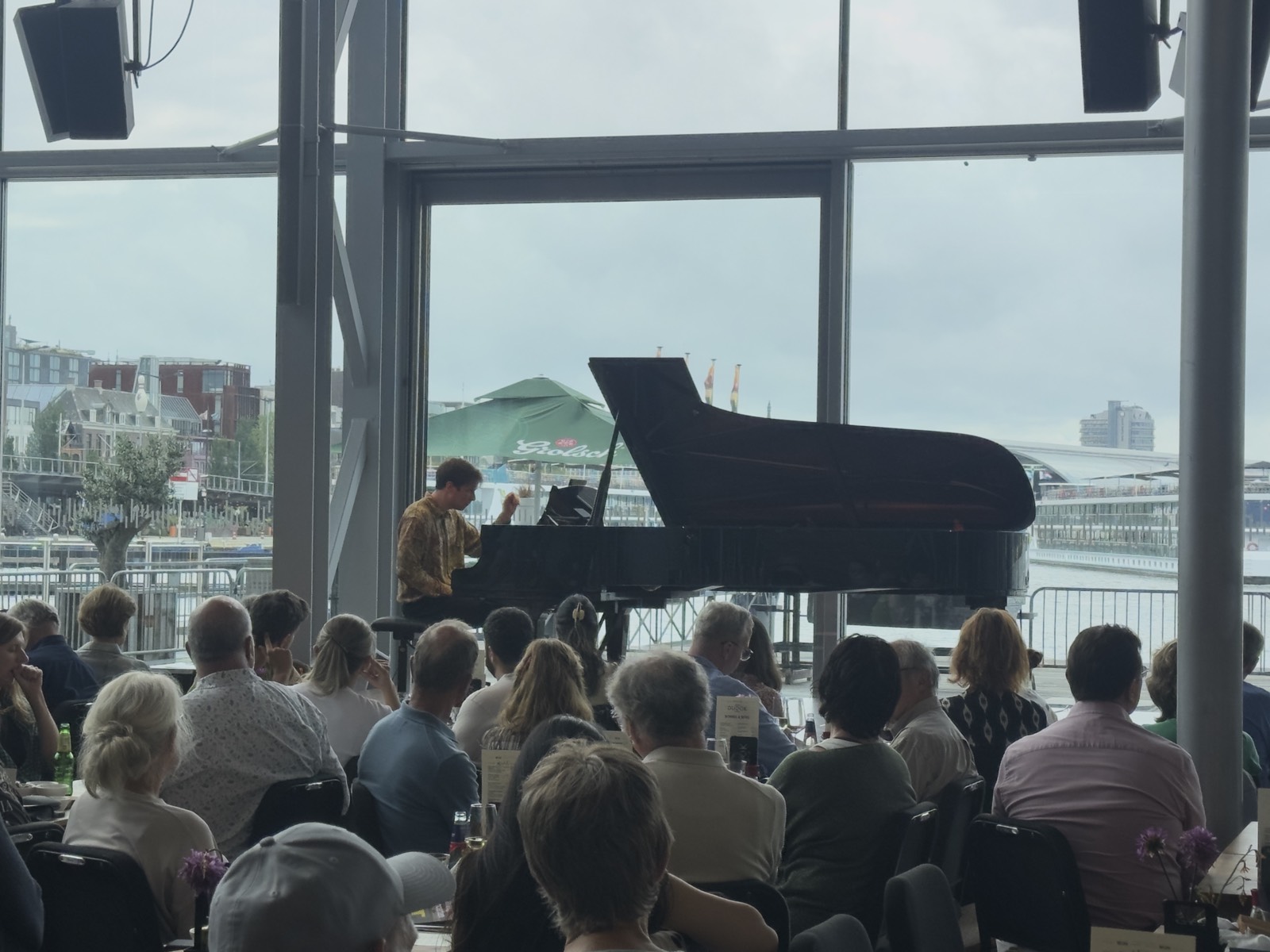Het publiek luistert in de Entreehal van het Muziekgebouw, met zicht op het IJ door de grote ramen