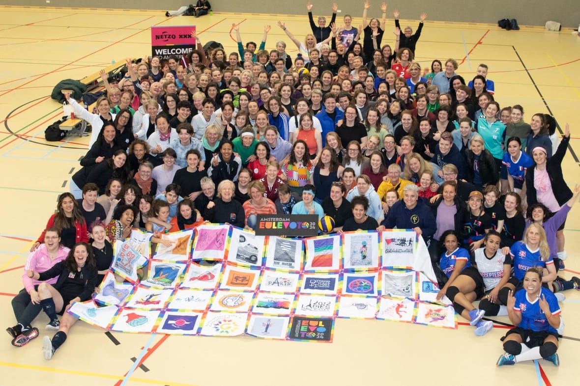Een grote groep deelnemers van het internationale volleybaltoernooi EuLeVoTo 2024 poseert in een sporthal, met een Netzo-welkomstbord op de achtergrond