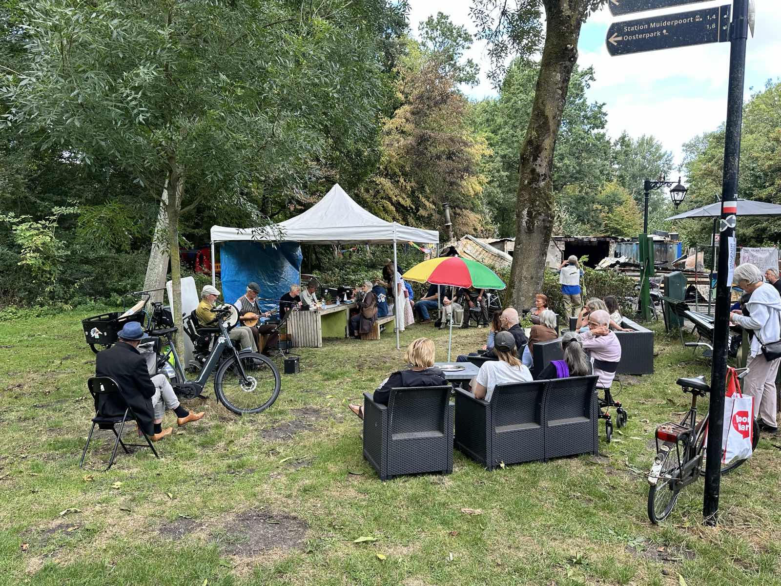 Bezoekers luisteren naar live muziek op het terrein van Parknest, met de uitgebrande resten op de achtergrond