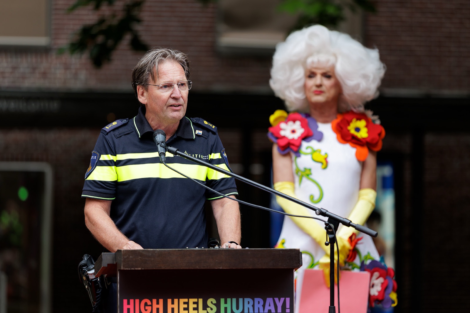 Politiechef Peter Holla spreekt de gasten toe vanachter het spreekgestoelte van High Heels Hurray, met Dolly Bellefleur op de achtergrond