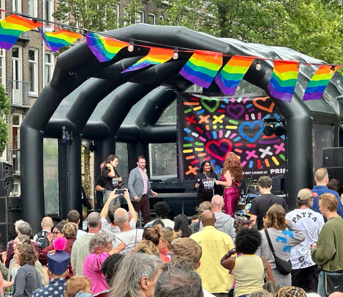 Een vol Javaplein voor het podium van Pride Oost, met regenboogvlaggen boven het publiek en de presentatie op het podium