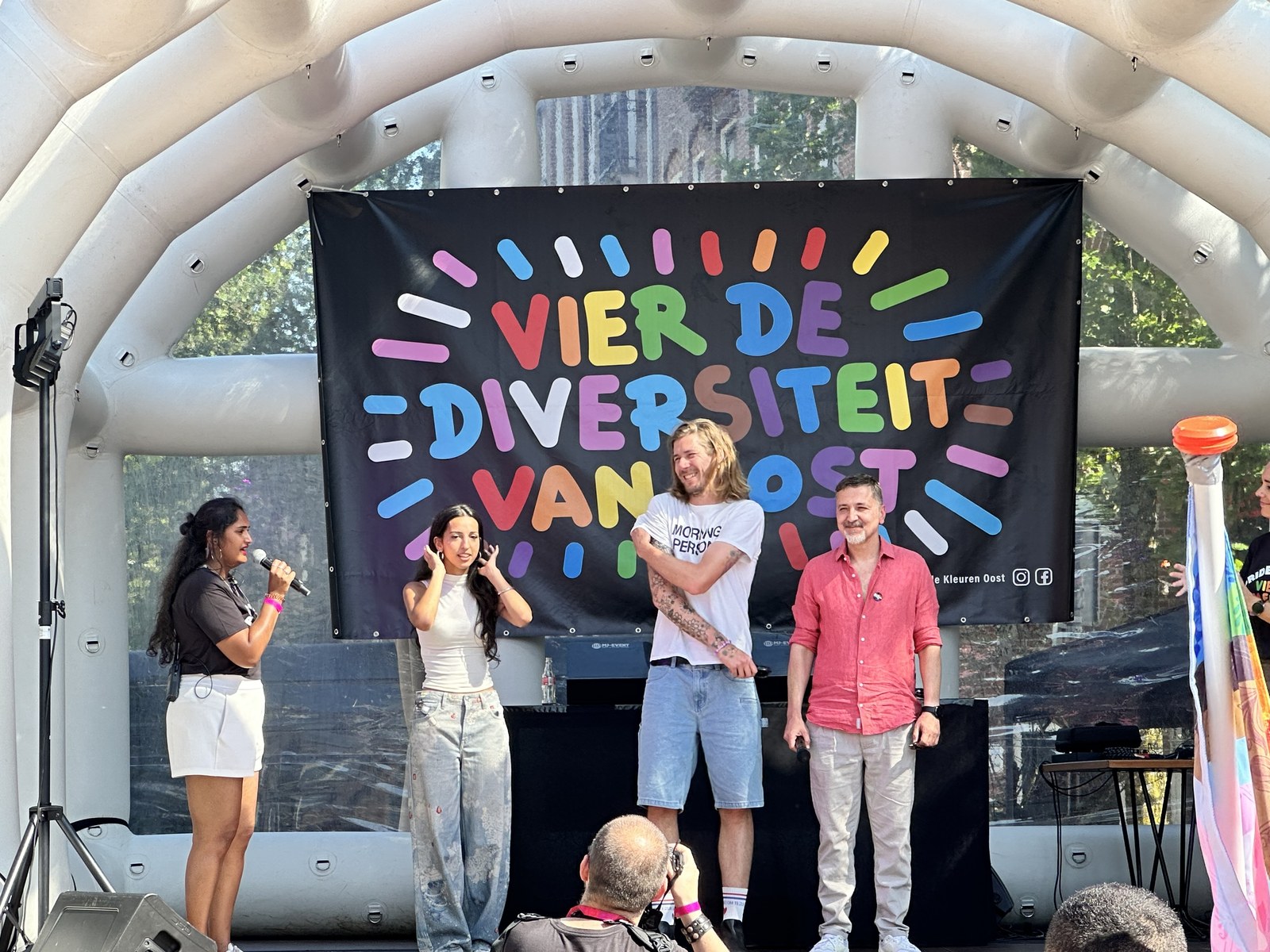 Jaika Koot ontvangt gasten op het podium tijdens de opening van Pride Oost, voor een banner met de tekst ‘Vier de diversiteit van Oost’
