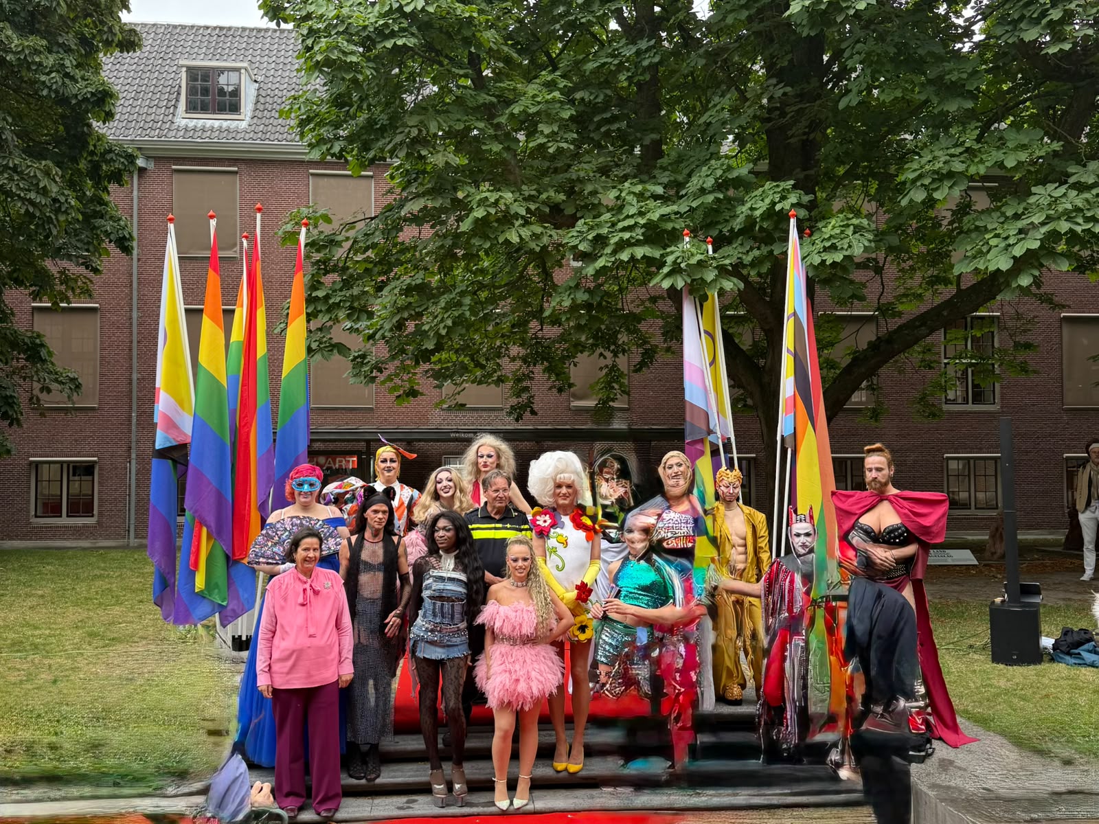 Groepsfoto met onder anderen Jan Pieter de Lugt, de dragqueens en dragkings en de regenboogvlaggen in de binnentuin van het H’ART Museum