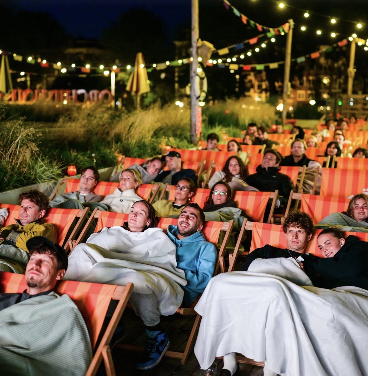 Volle rijen ligstoelen op het dakterras tijdens een avondvoorstelling, met lichtjes en vlaggetjes erboven