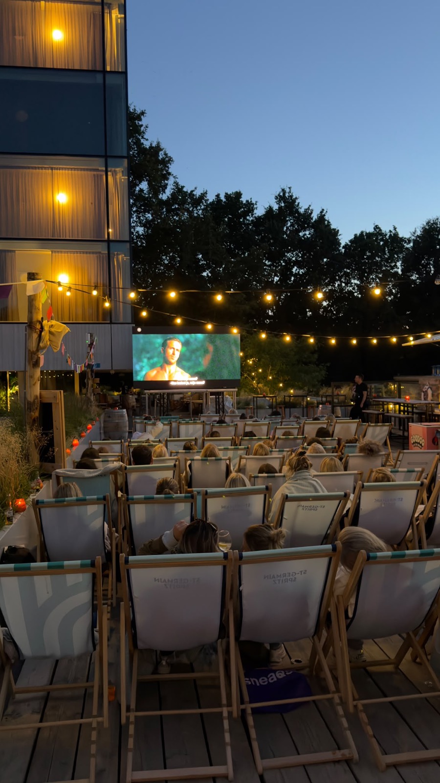 Het filmscherm van Strand van Oost in de schemering, met het dakterras en de lichtjes op de achtergrond