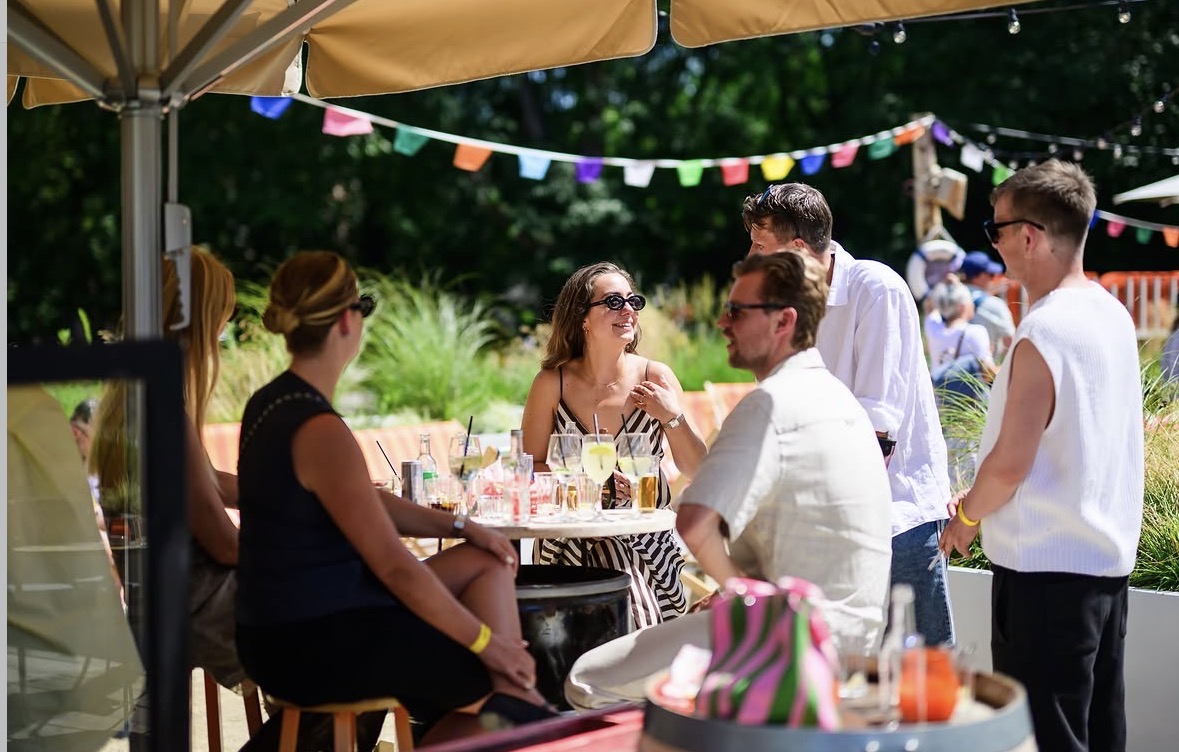 Gasten aan tafels op het zonnige terras van Strand van Oost, onder parasols en vlaggetjes