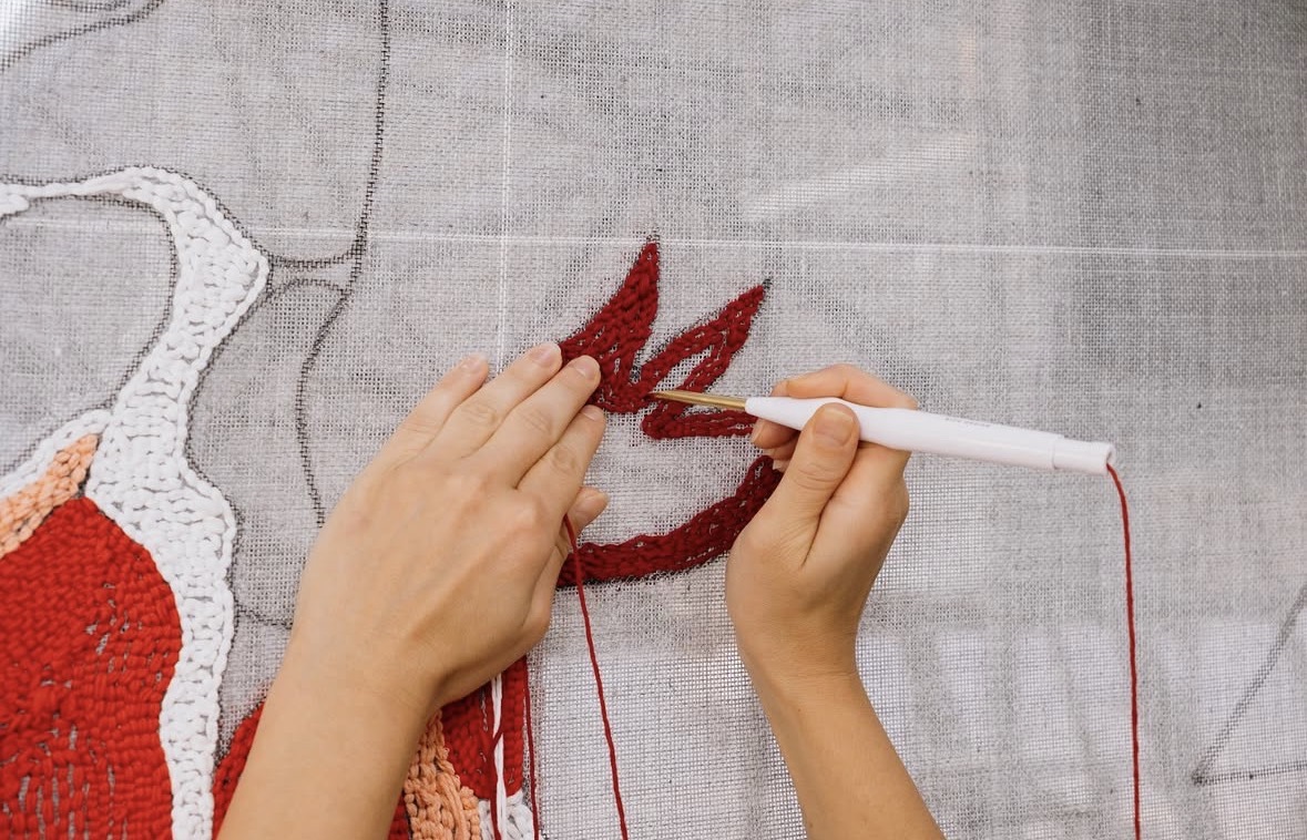 Twee handen werken met een punchnaald rood garen in het linnen doek
