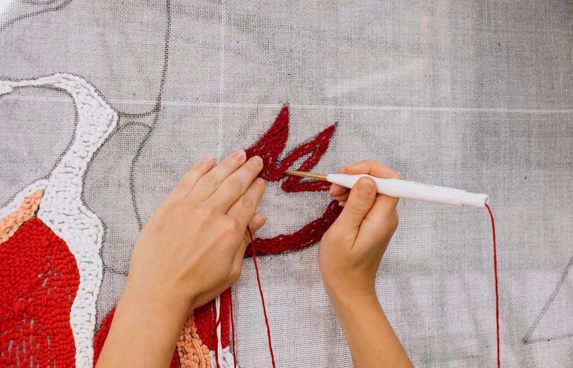 Twee handen werken met een punchnaald rood garen in het linnen doek