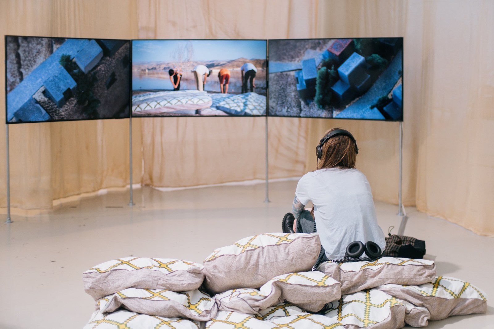 Een bezoeker bekijkt een video-installatie op drie schermen, gezeten op kussens